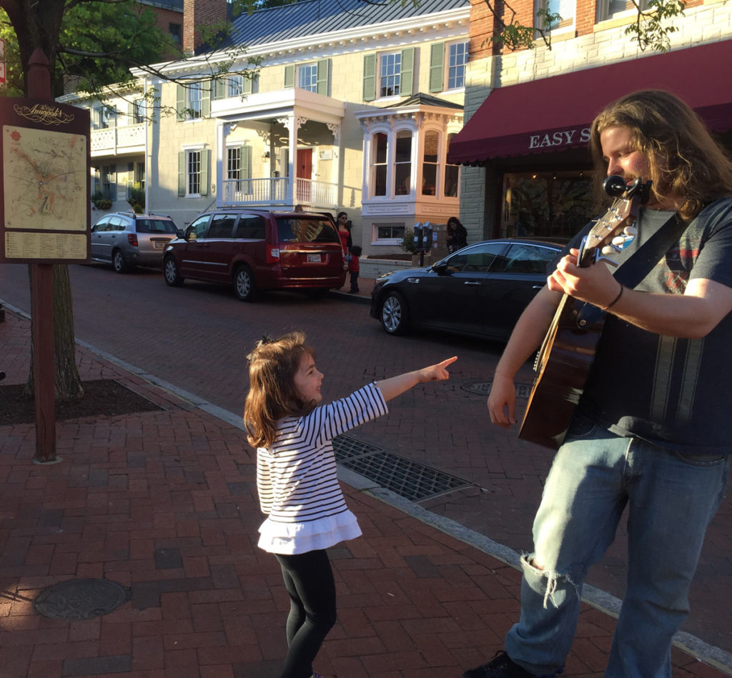 A little girl smiling and pointing to a man playing the guitr